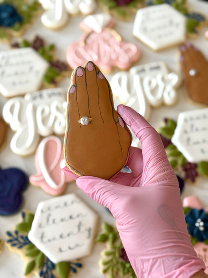 Hand holding a cookie shaped like a hand with a ring, surrounded by decorative cookies.