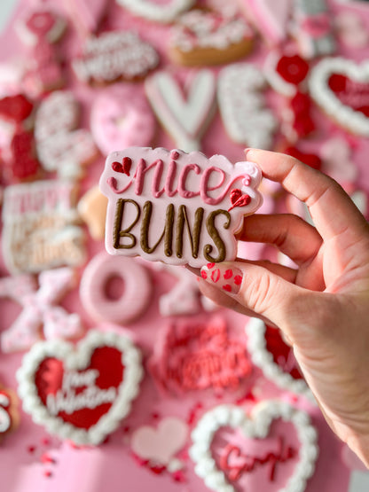 Hand holding a 'nice BUNS' cookie with pink and red decorative cookies in the background.
